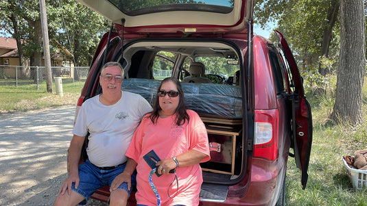 Two people sitting in the open trunk of a red dodge caravan with a camper conversion