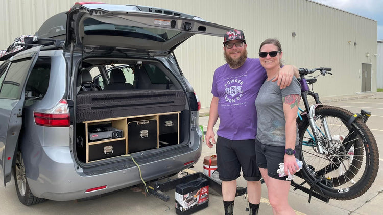 Two people with a bicycle on a car rack, standing next to an open Toyota sienna trunk with camper box.